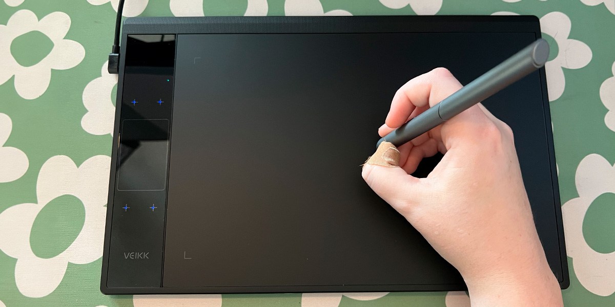 Hand using drawing pen on Veikk A30 V2 Drawing Tablet placed on background with green and white flowers